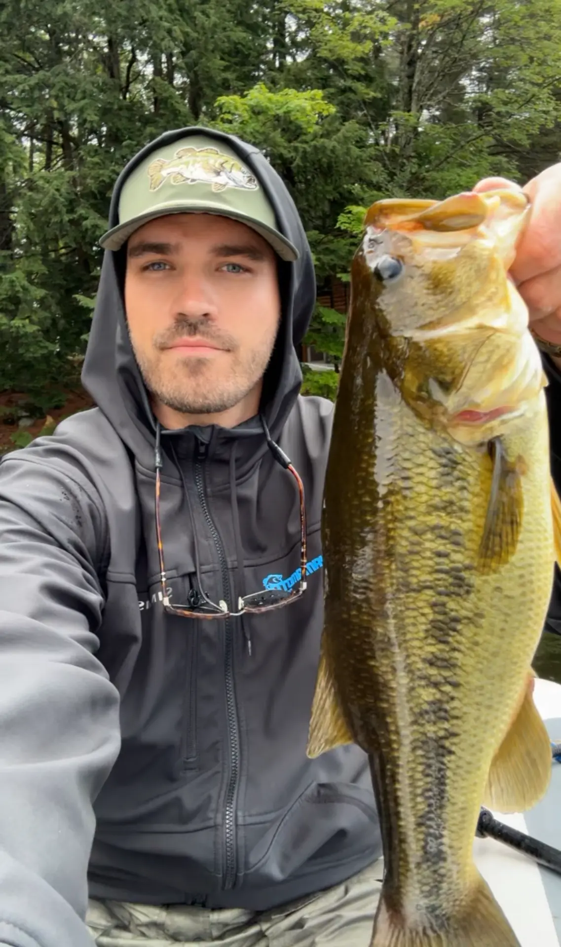 TJ LeClair holding a largemouth bass in a hooded jacket on the lake
